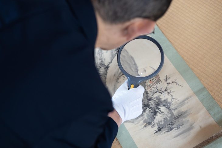 手袋をはめて虫眼鏡を持ち、掛け軸の状態を調べる人物