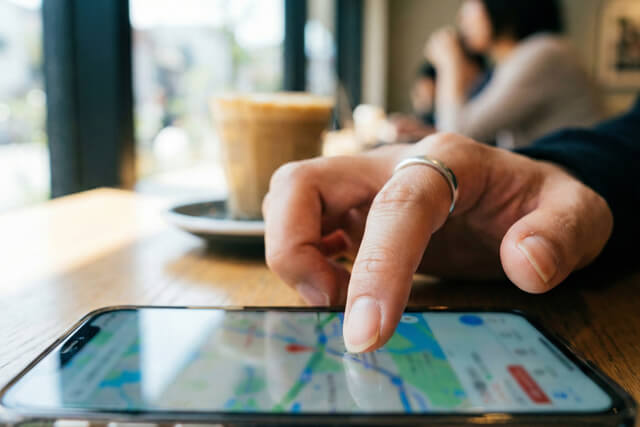 カフェでスマートフォンを使い、近くの貴金属買取店やリサイクルショップを地図アプリで検索している、指輪をした手元の写真