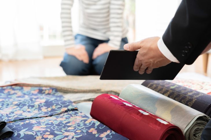 床に置かれた複数の反物の前に座っている人にタブレットの画面を見せる人の手元