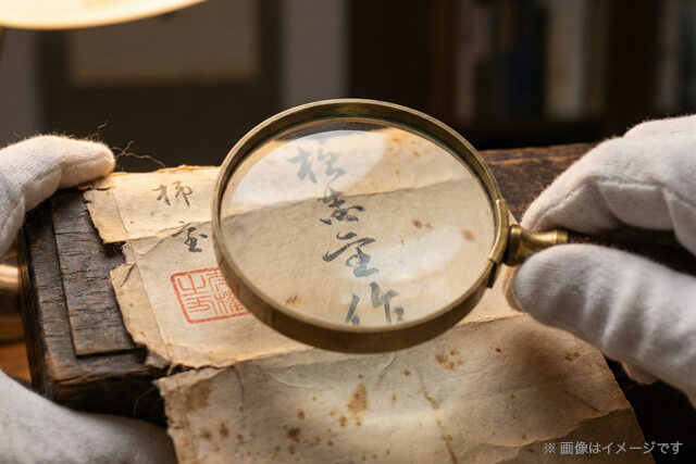プロの査定士がルーペを使って掛け軸の落款と共箱の箱書きを入念に確認している様子｜高額査定の重要ポイント