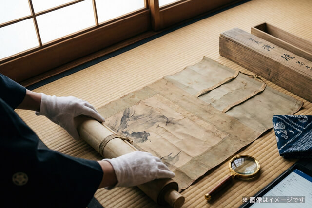 畳の上で白い手袋をしたプロの査定士が掛け軸を丁寧に査定している様子｜出張買取のイメージ画像