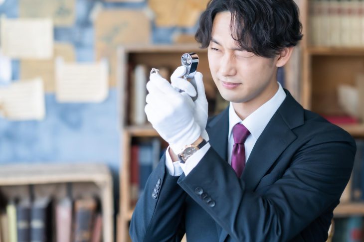 白い手袋をはめて持っているものをルーペで観察している男性の写真