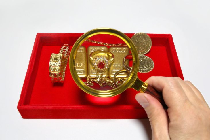 赤いトレイの上に置かれた金貨や貴金属をルーペで見る手元