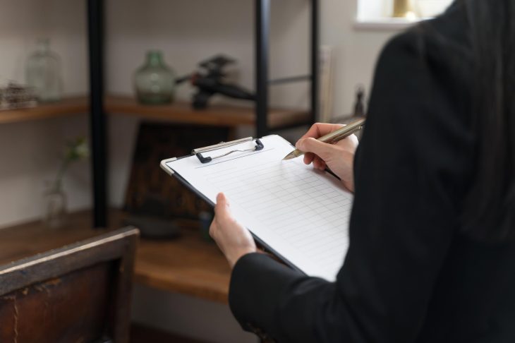 和風の部屋で、クリップボードに挟んだ紙にペンで書き込んでいるスーツを着た男性の手元