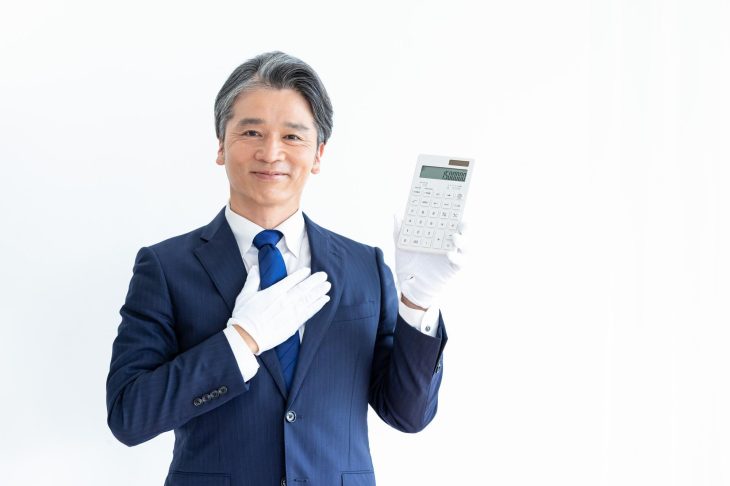 スーツとネクタイ姿の男性が白い手袋をはめて片手で電卓を持っている