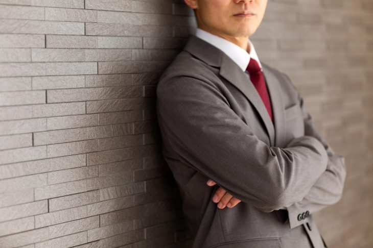 グレーのスーツにえんじのネクタイをしめ、腕を組んで壁にもたれかかっている男性の口元から上半身の写真
