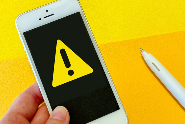 黄色い背景で、画面に警告(!)マークが表示されたスマートフォンを手に持つ