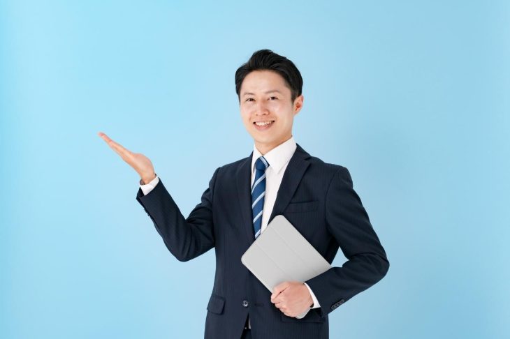 左手にタブレットを持ち右手の手のひらを上に向けているスーツ姿の男性