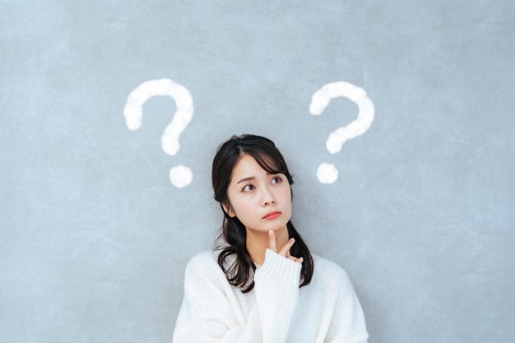 疑問符を背景に、顎に指を当てて考え込む女性