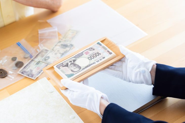古いコインや紙幣のそばで一万円札を載せたトレーを持つ人物の手元